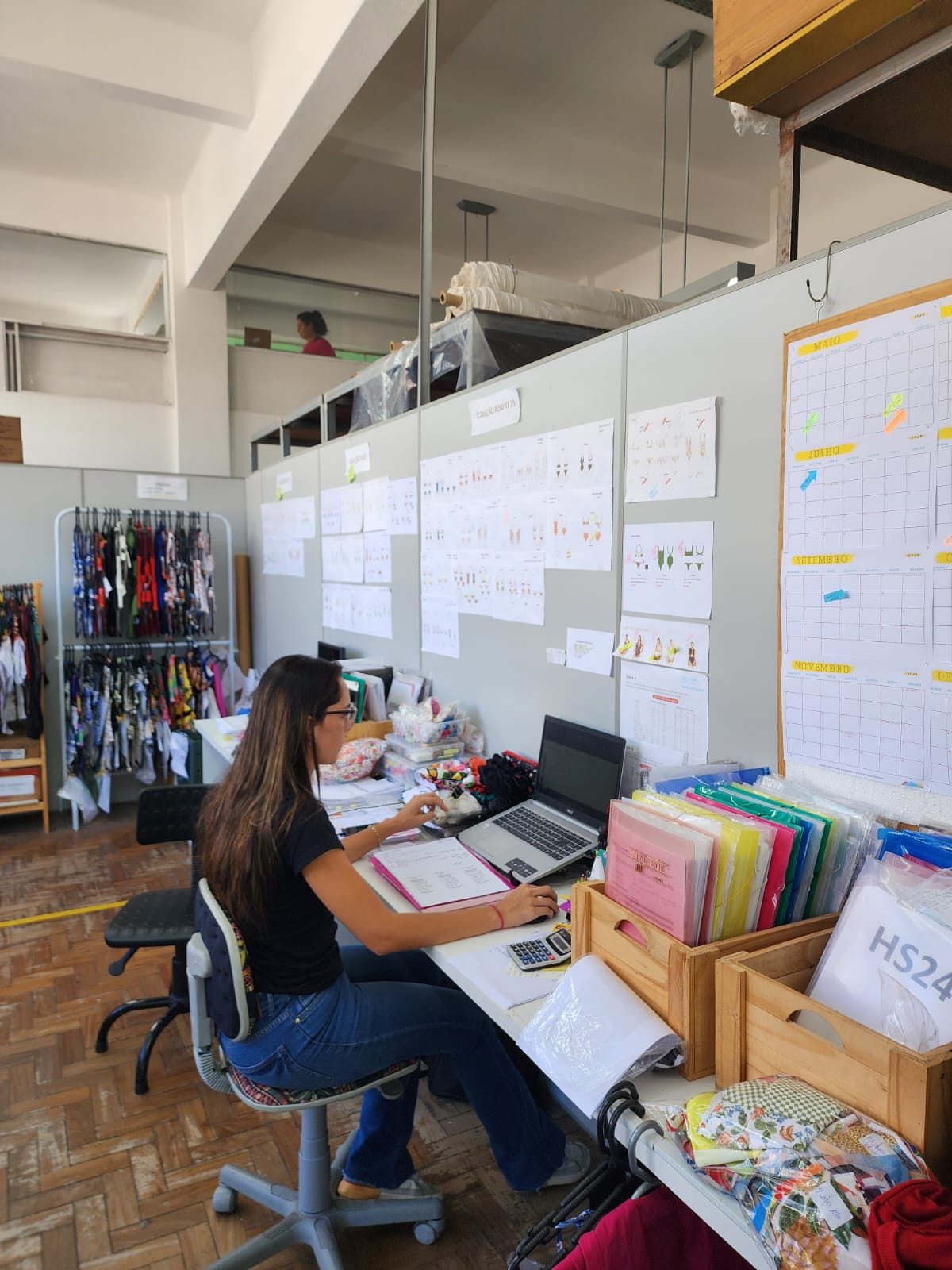 Colaboradora em sua mesa, com quadros de coleção e calendário de produção na parede à frente